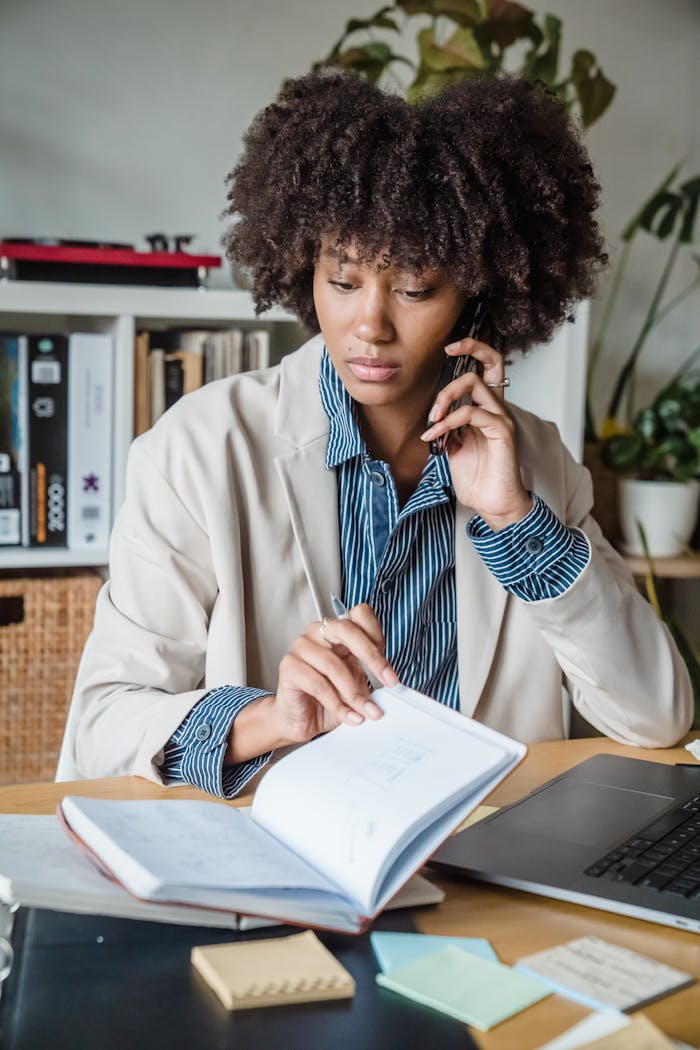 Focused woman multitasking in modern office with phone and notebook.